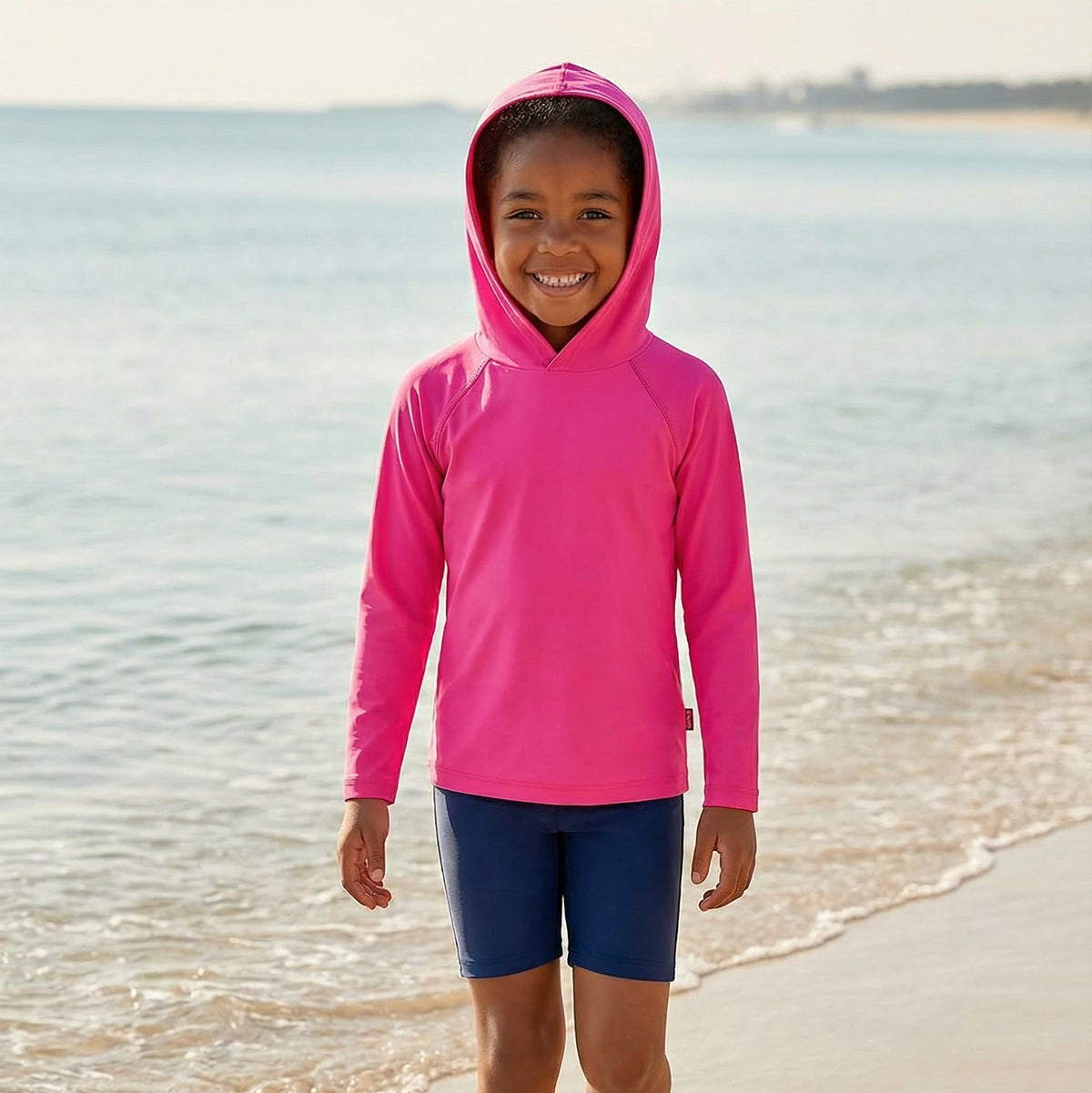 Child wearing a pink hoodie and blue shorts standing on a beach.