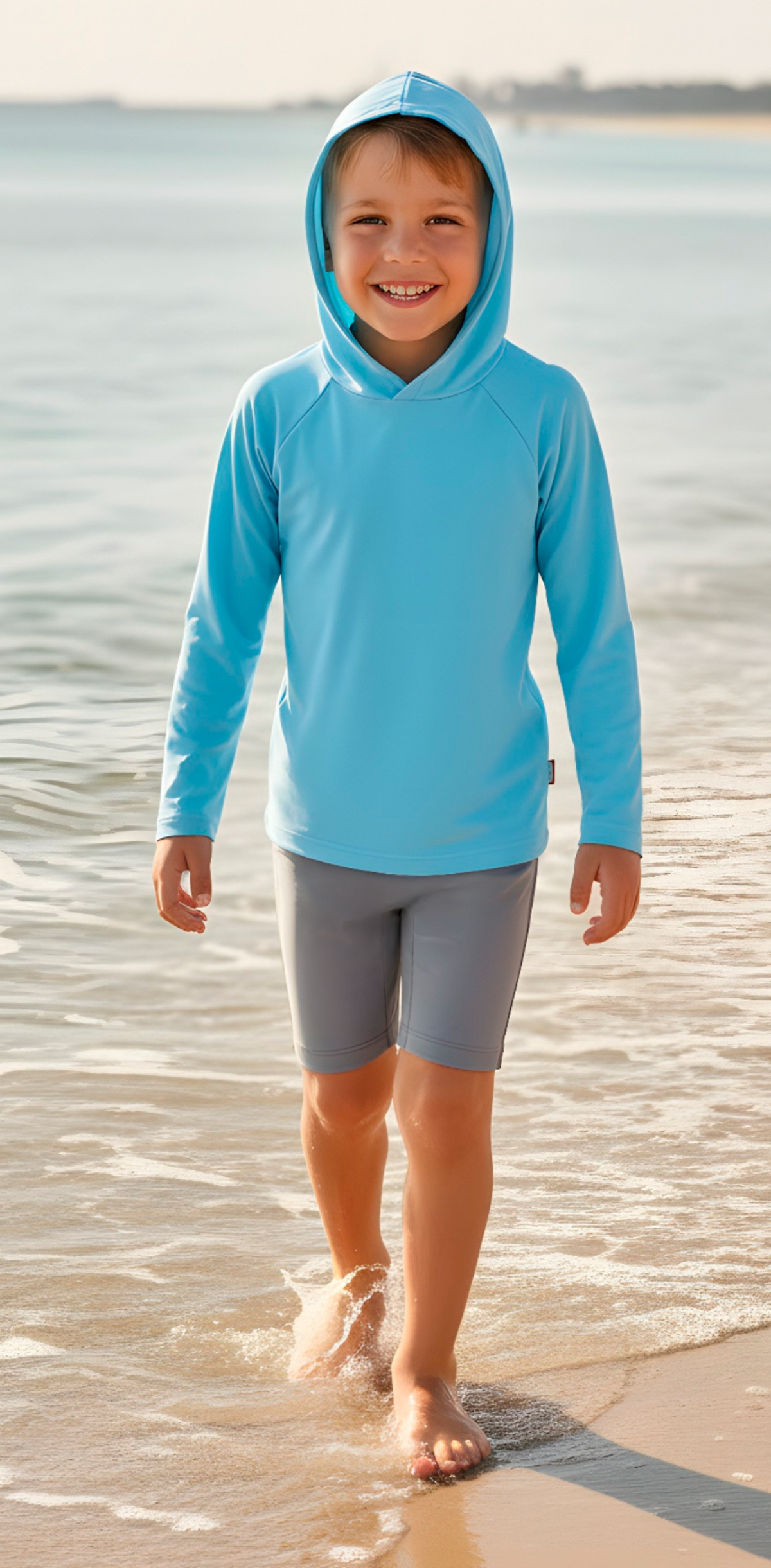 Child wearing a blue hoodie and gray shorts standing on a beach.