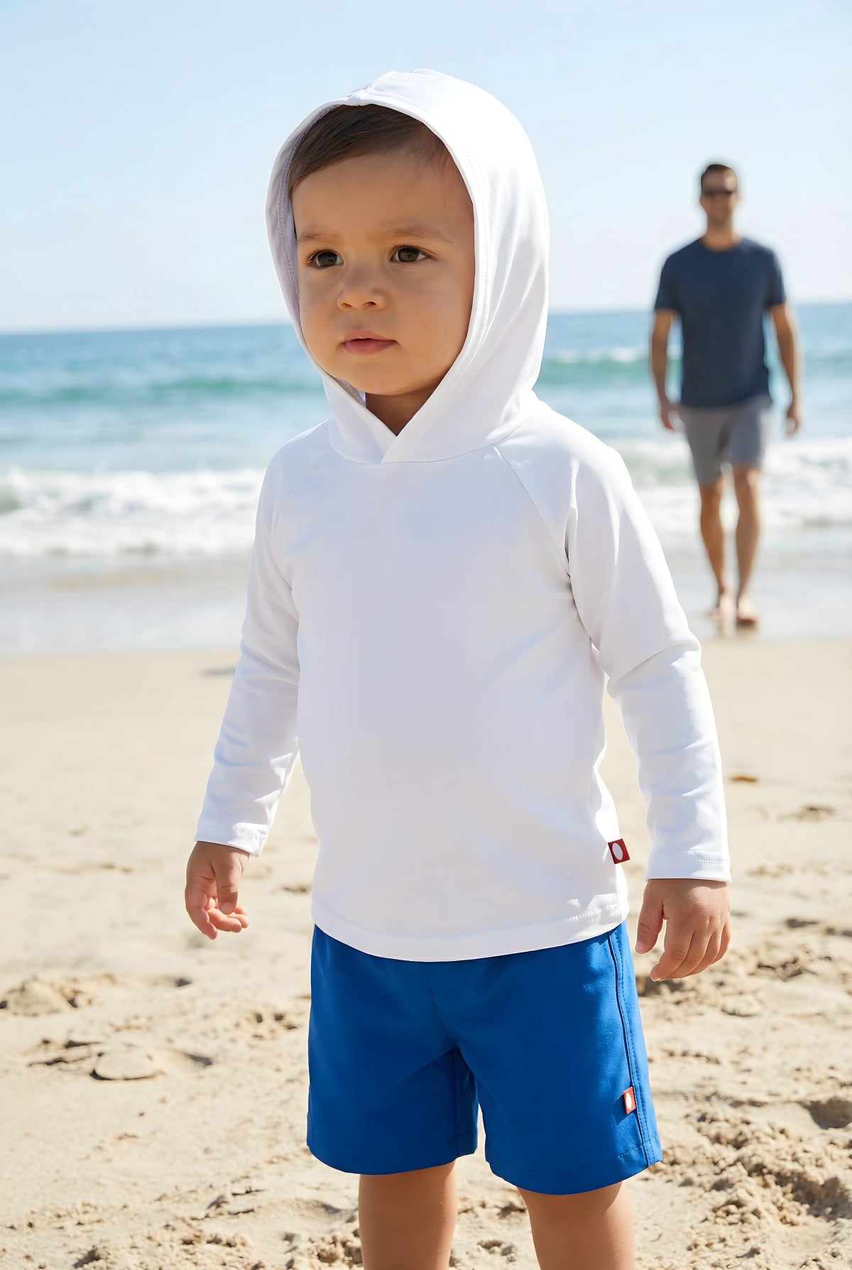 Child wearing a white hoodie and blue shorts on a beach with a person in the background.